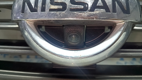 The grille assembly with the Nissan Quest 12-14 badge, camera, and molding is broken, worn, missing a fragment, broken fasteners, and peeling chrome.